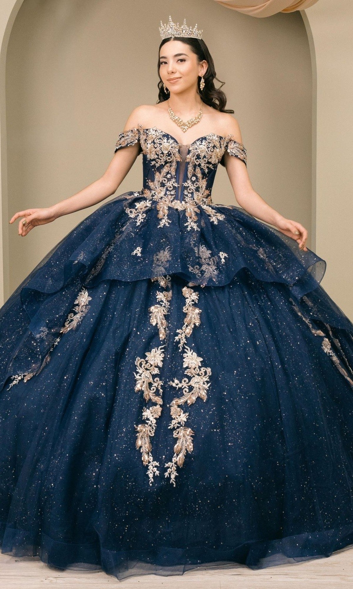 A woman poses elegantly against a beige background, wearing the Quinceanera Dress 8390JQ by Cinderella Couture in navy, adorned with intricate gold embroidery and complemented by a sparkling tiara.